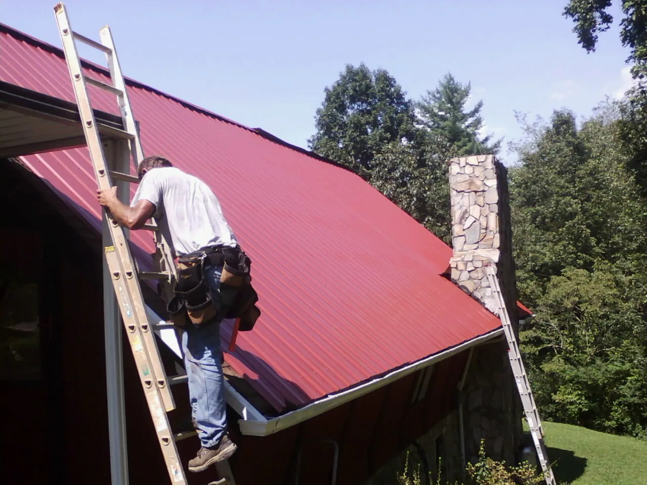 Roofer on a ladder beside a red metal roof and stone chimney — Soffit & Fascia Repair by Roofing Brothers in Country Club Hills